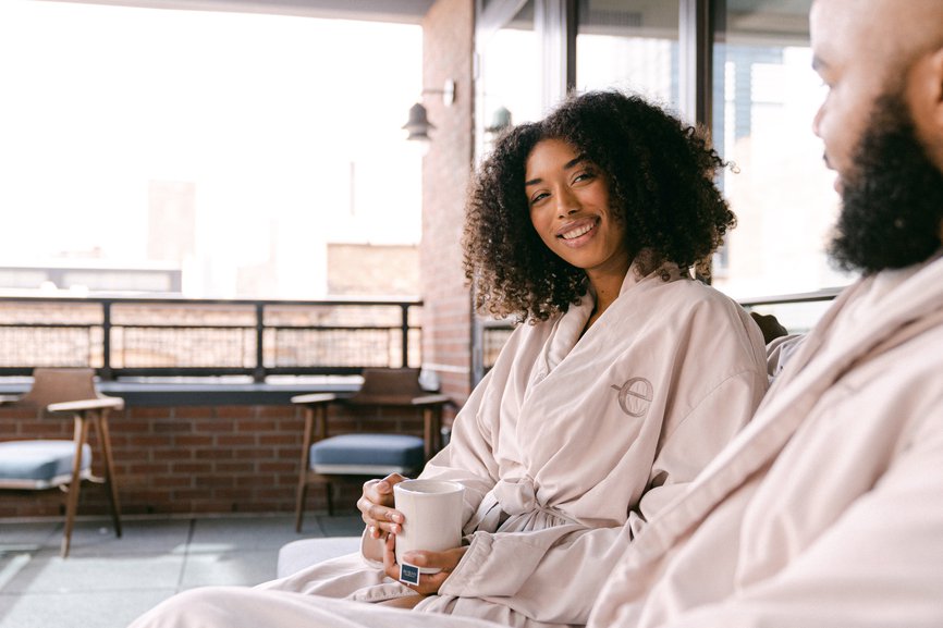 woman on a bed holding out a mug while coffee is poured into the mug