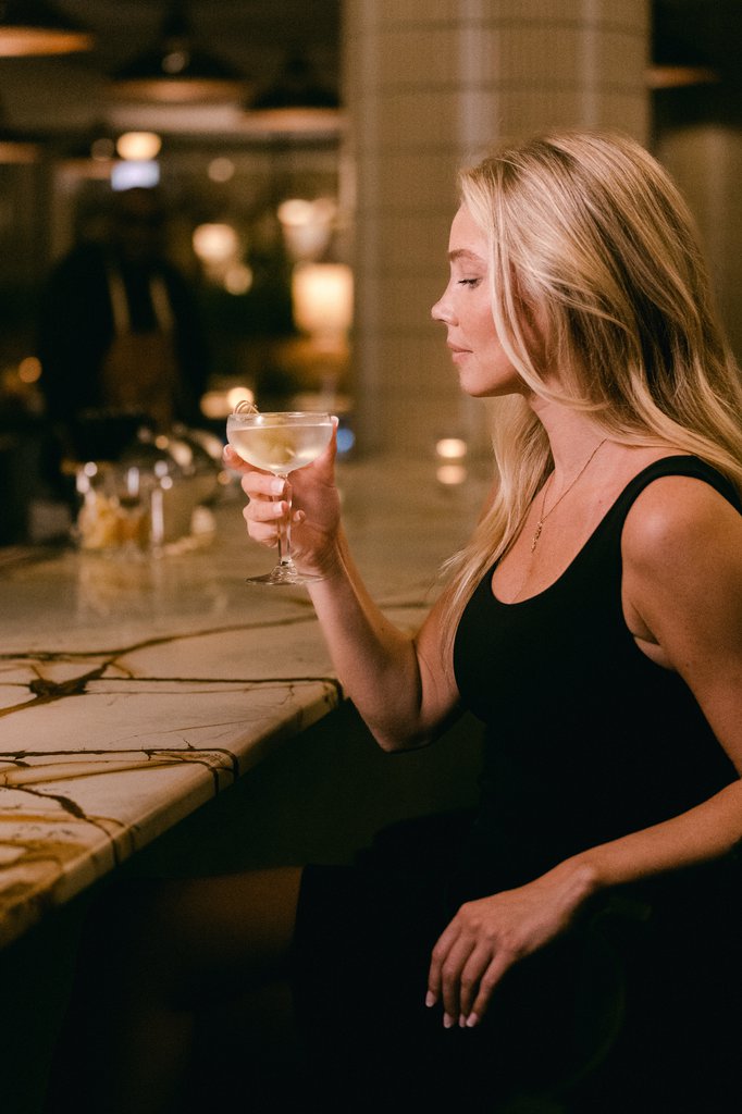 blonde hair woman sitting at the bar in black dress holding a glass of martini at fora chicago the emily hotel fulton market west loop