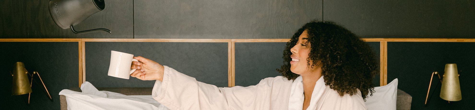 woman on a bed holding out a mug while coffee is poured into the mug