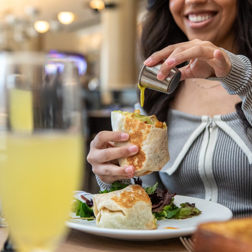 woman pouring sauce on breakfast burrito smiling , blurred mimosa in front